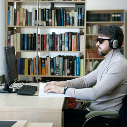 A man wearing headphones and glasses sits at a desk facing a computer and reading braille. There are shelves of books in the background. 