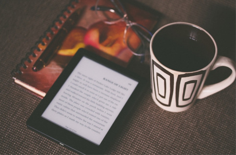 An e-reader resting on a notebook next to a pair of glasses and a cup of coffee
