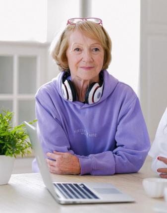 An elderly woman with headphones smiling in front of a computer