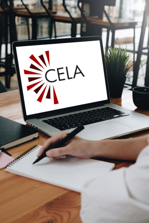 An open laptop on a desk displays the CELA logo. Beside it, a hand holds a pen poised over a notebook.