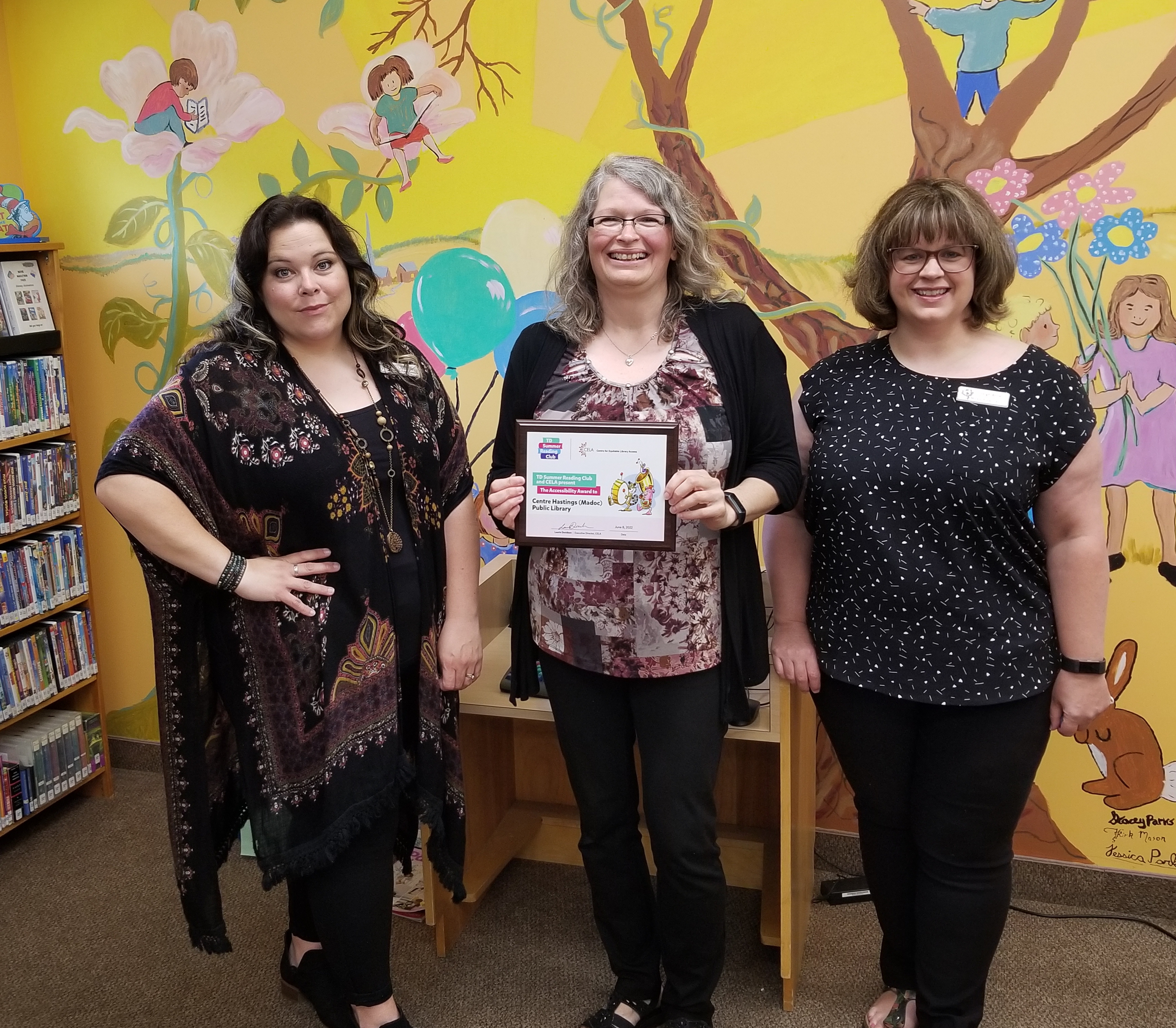 FlotsMembres du personnel de la bibliothèque publique de Centre Hastings tenant la plaque reçue dans le cadre du prix TDSRC. Ils se tiennent dans le département des enfants contre une peinture murale colorée.