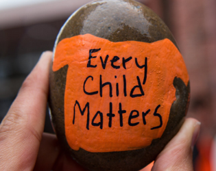 Hand holding a stone with an orange t-shirt painted on it. Written on the t-shirt in black are the words: &quot;Every Child Matters.&quot;