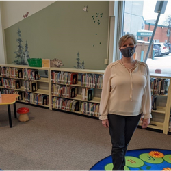 Une femme portant un masque se tient devant une rangée d'étagères basses dans une bibliothèque. Le mur et l'espace sont décorés de couleurs vives et de meubles pour enfants.