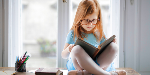 Red-headed child wearing glasses sits cross-legged on a table, reading a book.