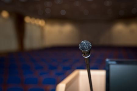 In the foreground is a microphone on a podium and the top left corner of a computer monitor. Out of focus in the background is an auditorium full of blue seats.