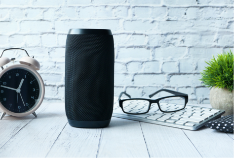 Un haut-parleur intelligent, une petite horloge, des lunettes et un clavier sur un bureau.