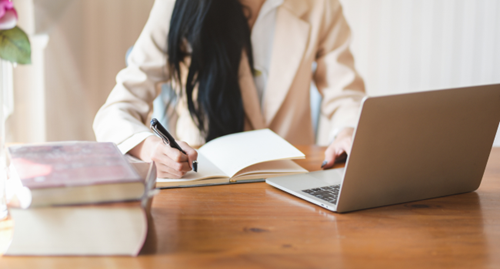 Le ventre d'une personne est visible derrière un bureau sur lequel elle écrit dans un cahier. Sur le bureau, il y a aussi un ordinateur portable ouvert et deux livres empilés.