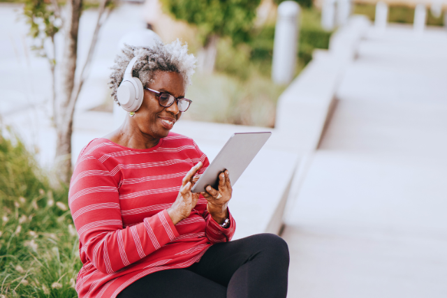 A person wearing glasses and headphones sits outdoors on a bench and holds a tablet device.