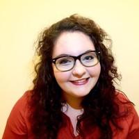 portrait of a smiling young woman wearing a reddish brown top, glasses and long dark curly hair. 