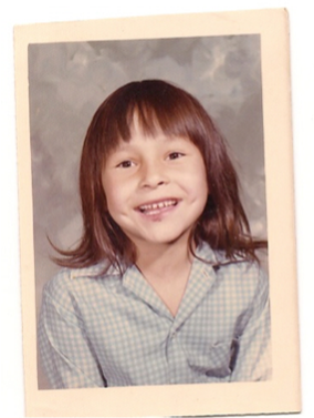 School portrait of Phyllis Webstad as a young girl. She has brown shoulder length hair with bangs and a grey shirt. She is smiling. 