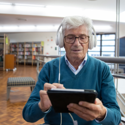 Older man is sitting in a library, listening to headphones while holding a tablet.