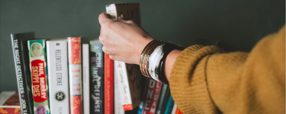 A woman wearing a gold sweater and a silver bracelet selecting a book from a bookshelf