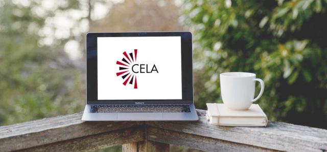 Laptop with black and white CELA logo on screen rests next to a mug and a book on a wooden balcony ledge
