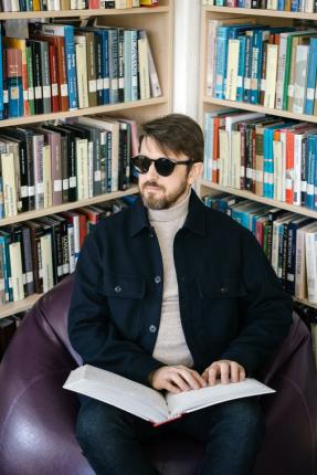 A man in a navy jacket reading braille while seated in front of a bookcase