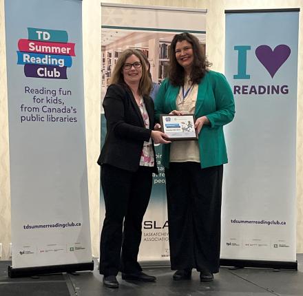 Two smiling women standing in front of TD Summer Reading Club banner holding the 2023 Accessibility Award plaque. One woman is taller with long brown hair and the other woman has light brown hair and wears glasses.