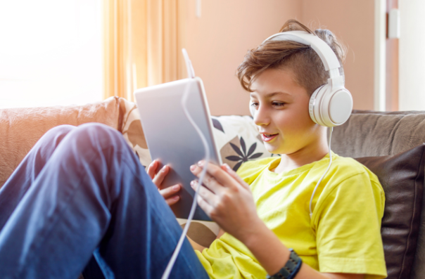 A child reclining on a couch while wearing headphones and looking at a tablet.