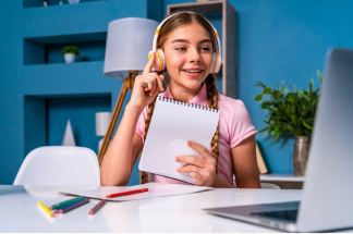 A child sitting in front of a  laptop. She is wearing headphones and holding a notebook.