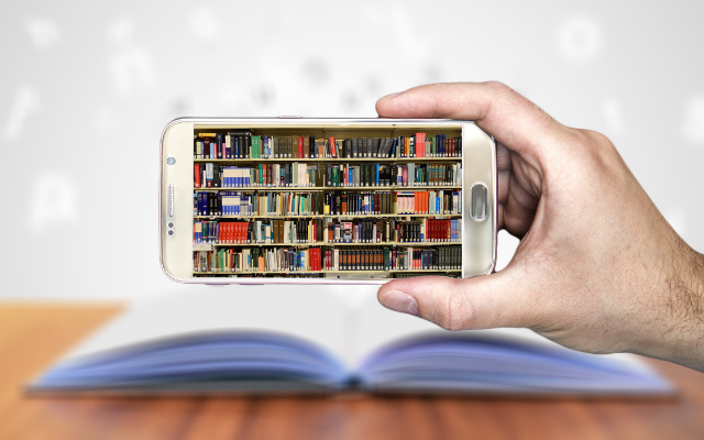 A hand holds a smartphone showing an image of bookshelves on the screen, a print book is open in background